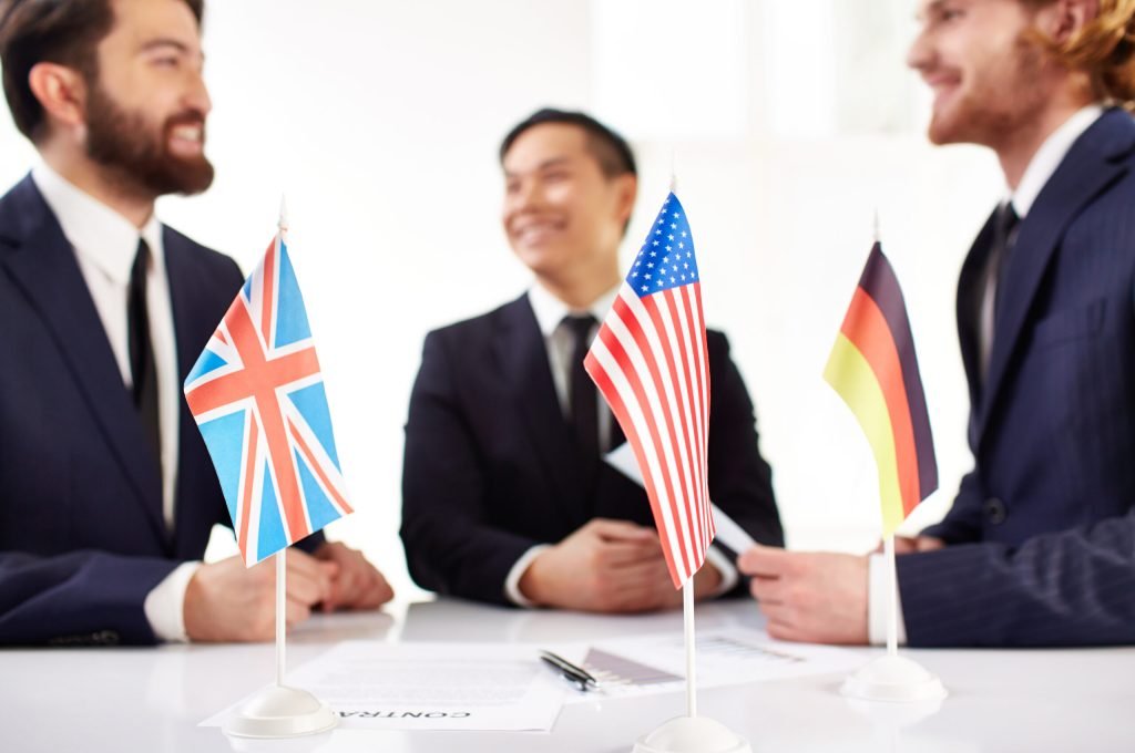 Image of American, British and German flags and business documents on workplace with three partners negotiating on background